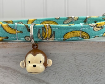 May include: A turquoise blue dog collar with a yellow buckle and a pattern of yellow bananas. A small monkey-shaped bell charm is attached to the collar.
