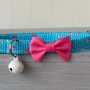 May include: A blue and white patterned cat collar with a pink bow and a white bell.