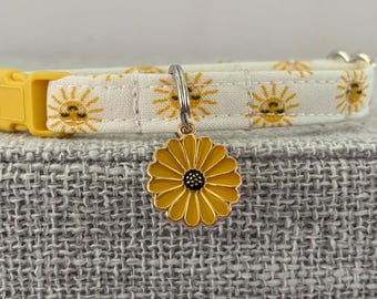 May include: A white fabric collar featuring a repeating yellow sun pattern with a yellow plastic buckle and a silver metal ring holding a yellow and black flower-shaped charm. The collar is designed with a secure clasp for easy adjustment and wear.