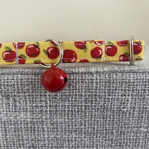 May include: A yellow cat collar with a red apple pattern and a red bell. The collar is made of fabric and has a metal D-ring and a yellow plastic buckle. The collar is approximately 1 inch wide.