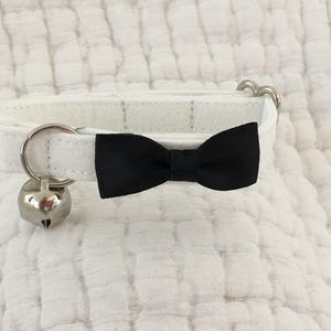 May include: White cat collar with a black bow tie and a silver bell. The collar is made of soft fabric and has a simple design.