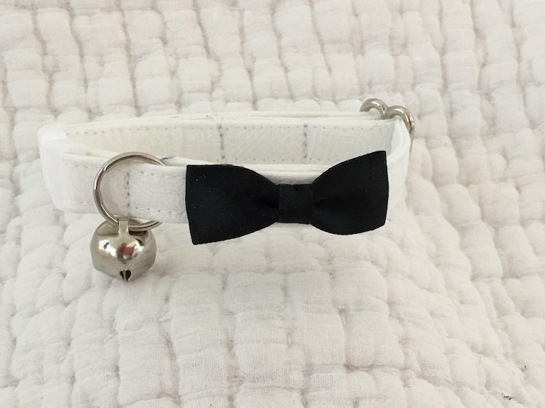 May include: White cat collar with a black bow tie and a silver bell. The collar is made of soft fabric and has a simple design.