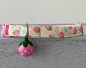 Pode incluir: Um colar de gato de tecido branco e rosa com estampa de morangos e um pingente de morango rosa e verde em forma de sino.