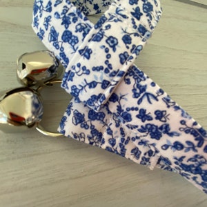 May include: A white and blue floral patterned ribbon with two silver bells attached.