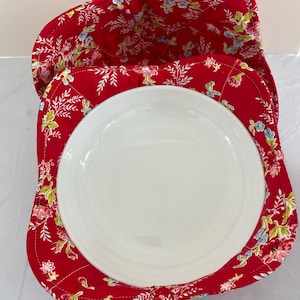 May include: A red bowl cozy with a floral pattern, holding a white ceramic plate. The cozy is made of quilted fabric with a design of pink, blue, and green flowers and leaves. The plate is round and has a smooth surface.