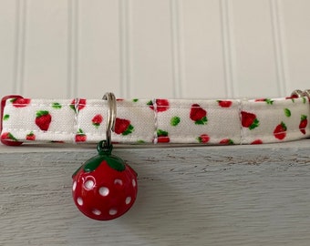 Pode incluir: Uma coleira branca para animais de estimação com um padrão de morangos vermelhos e uma campainha em forma de morango. A coleira tem uma fivela de metal e extremidades de plástico vermelho. A campainha é vermelha com bolinhas brancas e um caule verde.