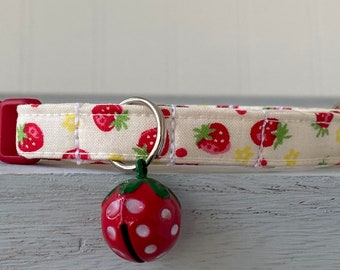 Pode incluir: Um colar de gato branco com uma fivela vermelha e uma estampa de morango. Um pequeno sino vermelho com bolinhas brancas pendura no colar.