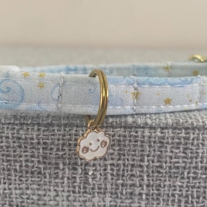 May include: A light blue cat collar with a white plastic buckle and gold-colored metal hardware. The fabric features a pattern of blue swirls and gold stars. A small cloud charm with a smiling face hangs from the collar.