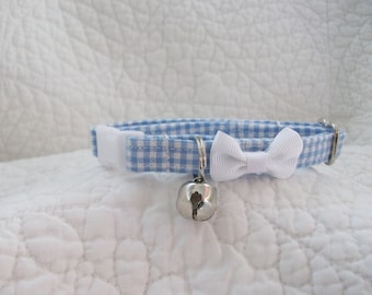 May include: A blue and white gingham cat collar with a white bow and a silver bell. The collar has a white plastic clasp and a metal loop for adjusting the size. The collar is approximately 0.5 inches wide.