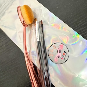 May include: A set of makeup brushes in a clear plastic bag. The brushes are black and rose gold. The bag has a holographic finish and a sticker with the text "Ole B" and "#becomebeautiful".