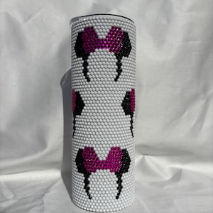 May include: A white tumbler covered in small, white rhinestones. The tumbler features a pattern of black and fuchsia mouse ears and bows. The design is repeated three times around the tumbler. The tumbler has a silver lid.
