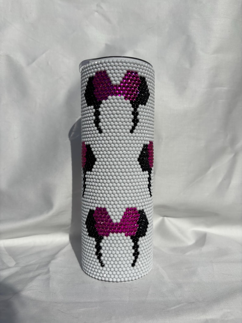 May include: A white tumbler covered in small, white rhinestones. The tumbler features a pattern of black and fuchsia mouse ears and bows. The design is repeated three times around the tumbler. The tumbler has a silver lid.