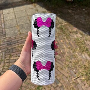 May include: White tumbler with a repeating pattern of black and pink Minnie Mouse bows and ears. The tumbler is covered in small, white, raised dots. The tumbler is cylindrical and has a silver lid.