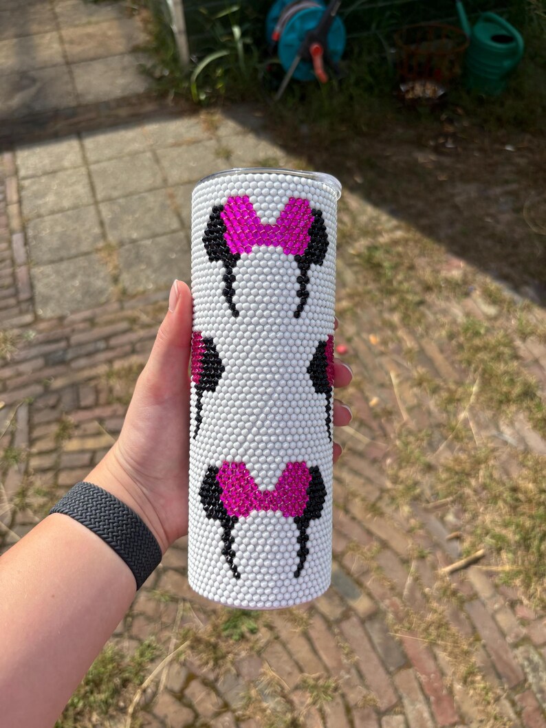 May include: White tumbler with a repeating pattern of black and pink Minnie Mouse bows and ears. The tumbler is covered in small, white, raised dots. The tumbler is cylindrical and has a silver lid.