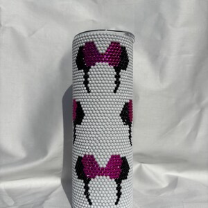 May include: A white tumbler covered in small, white rhinestones. The tumbler features a repeating pattern of black and pink bow-shaped designs. The tumbler is cylindrical and has a silver lid.