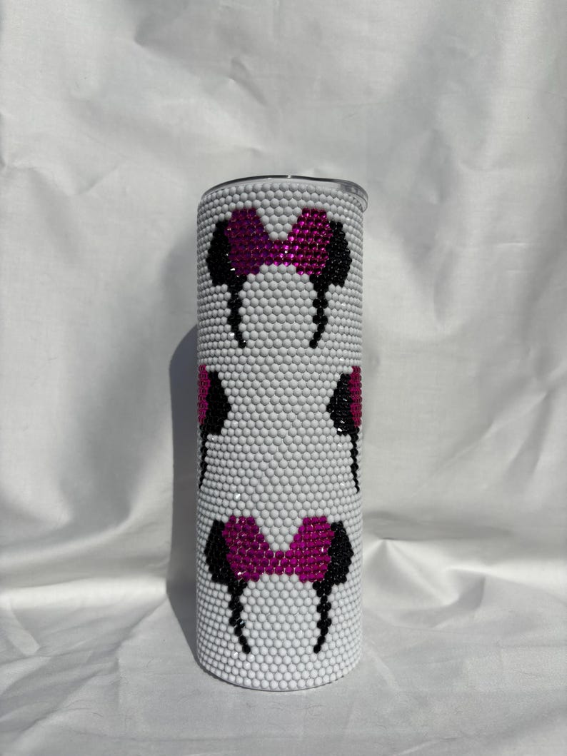 May include: A white tumbler covered in small, white rhinestones. The tumbler features a repeating pattern of black and pink bow-shaped designs. The tumbler is cylindrical and has a silver lid.
