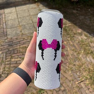 May include: White tumbler with a pattern of black and pink mouse ears and a pink bow. The tumbler is covered in small white beads. The design is repeated around the cylindrical shape of the tumbler.