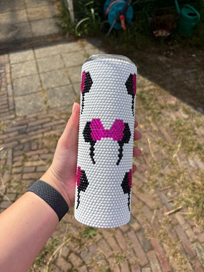 May include: White tumbler with a pattern of black and pink mouse ears and a pink bow. The tumbler is covered in small white beads. The design is repeated around the cylindrical shape of the tumbler.