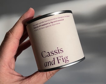 Puede incluir: Una pequeña lata de vela de plata con una etiqueta blanca. La etiqueta dice "Soy wax candle hand poured in Glasgow, Scotland Scents of raspberry, fig and coconut 105ml, 25 hour approx. burn time Cassis and Fig".