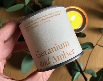 Puede incluir: Una lata de vela blanca con tapa plateada. La etiqueta está impresa con el texto "Soy wax candle hand poured in Glasgow, Scotland. Scents of geranium, amber and sandalwood.  Burn time 26 hour approx." en texto marrón. La vela está etiquetada como "Geranium and Amber" en letras marrones grandes.