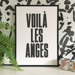 Puede incluir: Una impresión enmarcada en negro con el texto "VOILA LES ANGES" en letras blancas. La impresión está colgada en una pared con un patrón abstracto en blanco y negro. Dos pequeñas latas de metal con etiquetas están en primer plano.