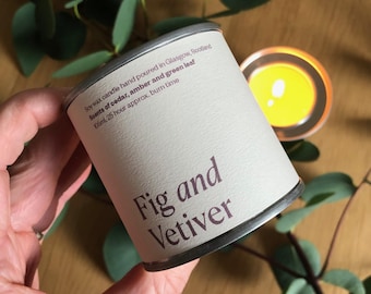 Puede incluir: Una vela de lata blanca con una etiqueta marrón que dice "Fig and Vetiver". La etiqueta también incluye el texto "Soy wax candle hand poured in Glasgow, Scotland. Scents of cedar, amber and green leaf. 106ml, 25 hour approx. burn time."