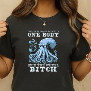 Könnte beinhalten: Dunkelgraues T-Shirt mit einem blauen Oktopus, der eine Pfeife raucht. Der Text lautet "I'M LIKE 12 PEOPLE PACKED IN ONE BODY SPIN THE WHEEL BITCH". Der Oktopus hat detaillierte Tentakel und einen strukturierten Kopf.