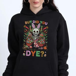 Puede incluir: Sudadera con capucha negra con un gráfico colorido. Presenta un esqueleto con orejas de conejo, rodeado de huevos de Pascua y un conejo rosa. El texto "BUT DID YOU DYE?" se muestra en colores vibrantes.