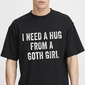May include: A black short-sleeved t-shirt featuring the white distressed-style text I NEED A HUG FROM A GOTH GIRL printed across the chest. The garment has a crew neckline and a standard fit, showcasing clear, bold lettering against the dark fabric.