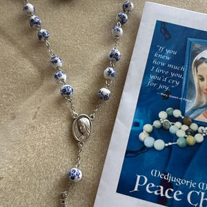 May include: A rosary featuring white ceramic beads with blue floral patterns, connected by silver-toned metal links. The rosary includes a silver-toned metal centerpiece depicting a face and a silver-toned crucifix at the bottom end.