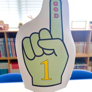 Peut inclure: Une main en mousse en forme de main avec l'index pointé vers le haut. La main est vert clair avec un contour blanc et le chiffre "1" en jaune. Le mot "GOD" est écrit verticalement sur l'index.