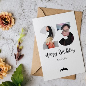 K&ouml;nnte beinhalten: Eine wei&szlig;e Geburtstagskarte mit der Zahl 23, die Fotoausschnitte einer Person enth&auml;lt, sowie dem Text Happy Birthday und AMELIA und einem kleinen schwarzen Kuchen-Symbol. Die Karte liegt auf einem braunen Umschlag mit pfirsichfarbenen Blumen.