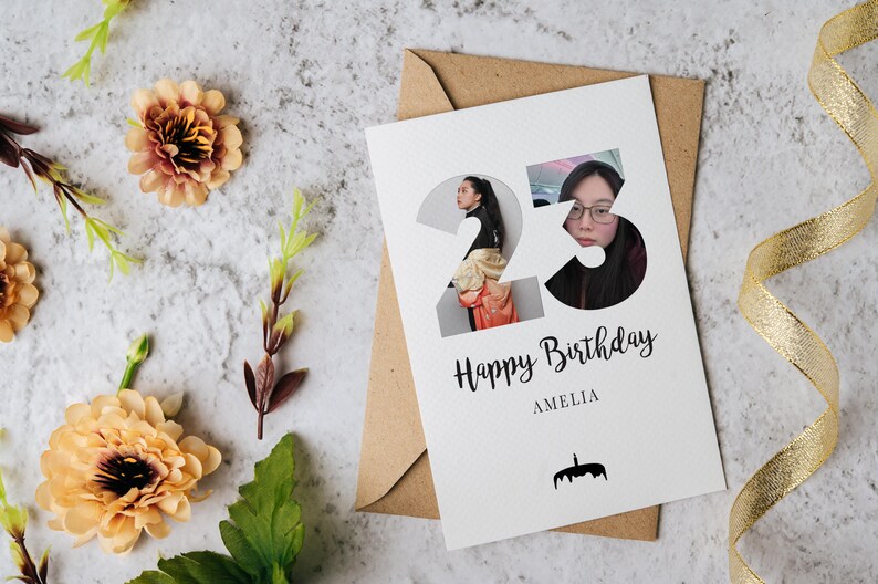 Może przedstawiać: Biała kartka urodzinowa z liczbą 23 zawierającą wycięcia ze zdjęciami osoby, napisem Happy Birthday i AMELIA oraz małą czarną ikoną tortu. Kartka umieszczona jest na brązowej kopercie z brzoskwiniowymi kwiatami i złotą wstążką.