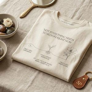 Puede incluir: Una camiseta color crema con el texto "NOT EVERYTHING NEEDS TO BLOOM RIGHT NOW" y una ilustración botánica. La imagen también presenta pequeños cuencos con piedras y flores secas, un pequeño vial de vidrio y un llavero de cuero.