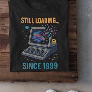 Op de afbeelding: Zwart t-shirt met een retro computer graphic en de tekst "STILL LOADING... SINCE 1999". Het ontwerp toont een vintage computer met een kleurrijk scherm en een smiley. Het shirt wordt gepresenteerd op een houten oppervlak.