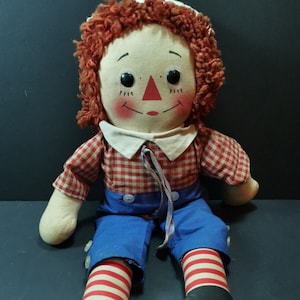 May include: A vintage rag doll with a fabric face, red yarn hair, and a red triangle nose. The doll wears a red and white checkered shirt, blue pants, and black shoes. Its legs have red and white striped socks.