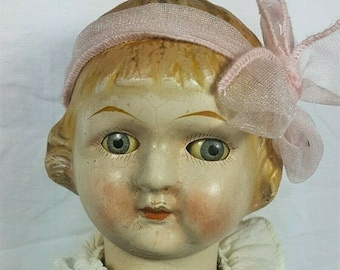 May include: Antique doll with a painted face, blue eyes, and a pink headband with a bow. The doll is wearing a white dress with a ruffled collar. The doll's skin has a cracked, aged appearance.