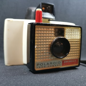 May include: A vintage white and black Polaroid Swinger Model Land Camera with a red flash bulb. The camera has a grid pattern on the front and the text "POLAROID LAND CAMERA SWINGER MODEL" is printed on the bottom.