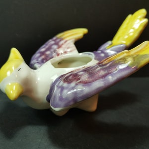 May include: A ceramic bird planter with yellow, purple, and white paint. The bird has its wings spread out and its head tilted to the side. The planter has a hole in the top for planting.