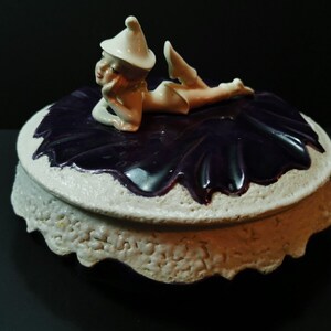 May include: A ceramic trinket box with a white and purple design. The lid features a white figurine of a fairy laying on a purple fabric.