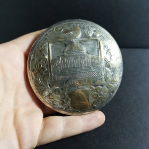 May include: Silver and gold toned round compact with an engraved image of the Dome of the Rock in Jerusalem. The compact has a floral design around the edge.