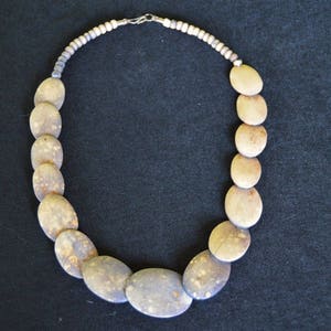 May include: A necklace featuring large, oval-shaped, beige and gray stones. The stones are strung together with a smaller, beaded strand and a silver-colored clasp. The necklace is displayed against a dark background.