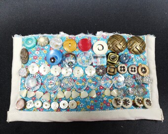May include: A blue fabric panel with a white border is covered in a variety of buttons. The buttons are in various colors, shapes, and sizes, including white, blue, red, yellow, and brown. Some buttons have a floral pattern.