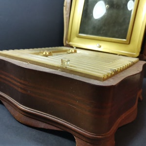 Puede incluir: Una caja de madera para joyas con una tapa con bisagras y un espejo en el interior. La caja tiene un acabado marr&oacute;n con un borde dorado y un dise&ntilde;o decorativo en la tapa.