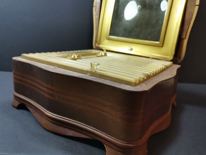 Puede incluir: Una caja de madera para joyas con una tapa con bisagras y un espejo en el interior. La caja tiene un acabado marr&oacute;n con un borde dorado y un dise&ntilde;o decorativo en la tapa.