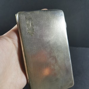 May include: A silver cigarette case with a textured surface and a monogrammed "SG" on the front.