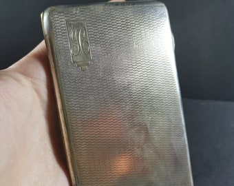 May include: A silver cigarette case with a textured surface and a monogrammed "SG" on the front.