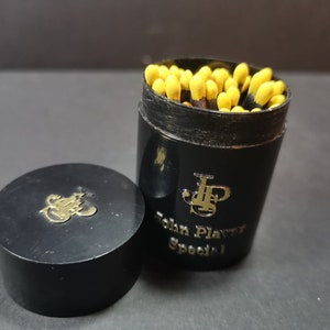 May include: A black round container with a gold John Player Special logo and a lid. The container is filled with yellow matches.