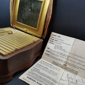 Puede incluir: Un radio y fon&oacute;grafo Emerson vintage, modelo 570 Executive Desk Set, en un gabinete de madera con una rejilla de altavoz dorada. La radio tiene una antena incorporada y funciona con bater&iacute;as. Las instrucciones de la radio son visibles en la imagen.