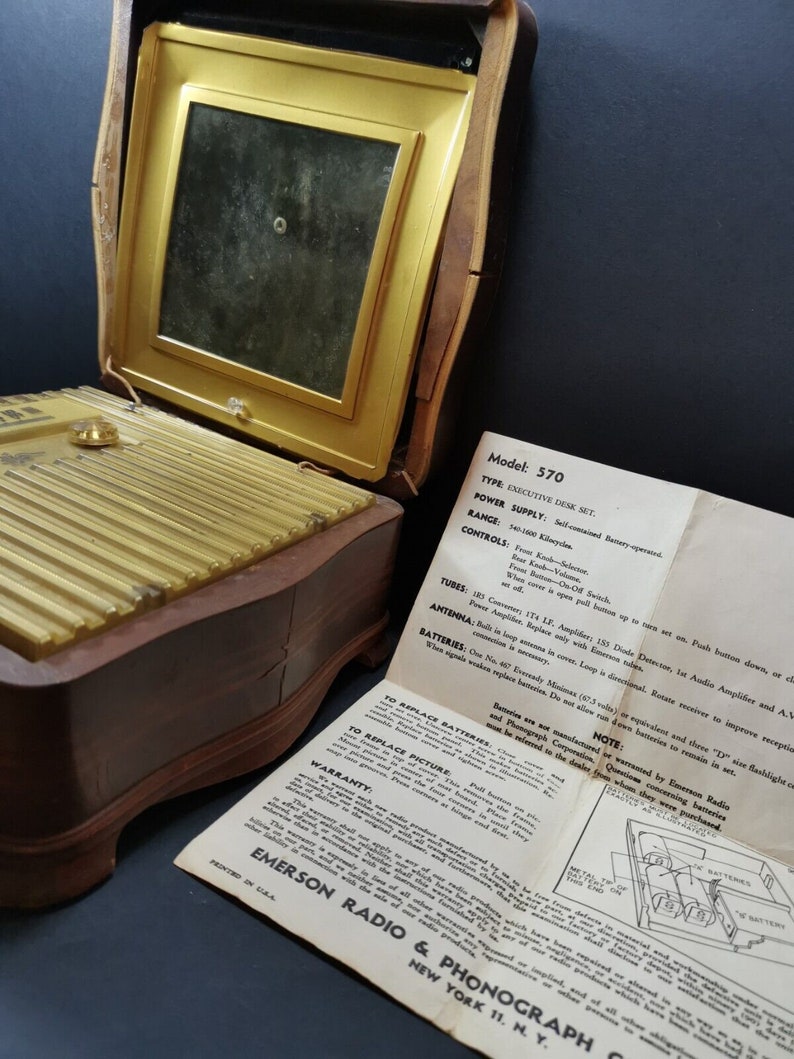 Puede incluir: Un radio y fon&oacute;grafo Emerson vintage, modelo 570 Executive Desk Set, en un gabinete de madera con una rejilla de altavoz dorada. La radio tiene una antena incorporada y funciona con bater&iacute;as. Las instrucciones de la radio son visibles en la imagen.